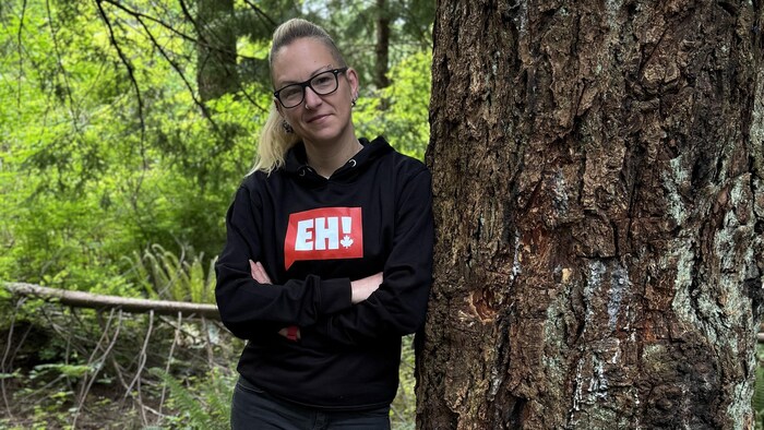 Jessica Glowacki a les bras croisés et sourit subtilement en regardant devant elle, appuyée sur un arbre.