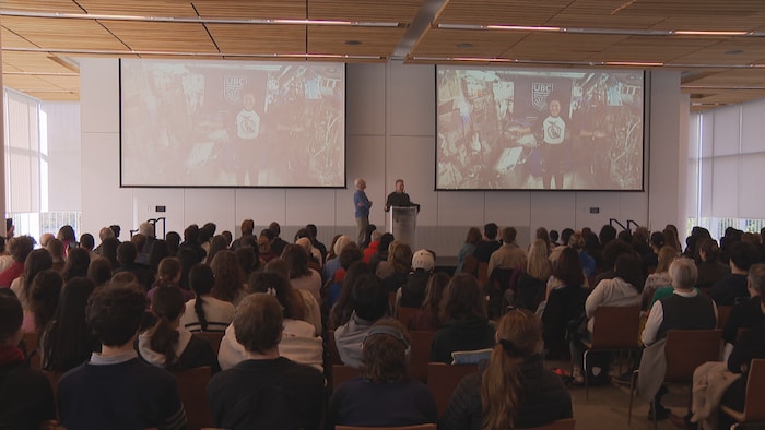 Une grande salle de conférence remplie de personnes regardant deux écrans sur lesquels est projeté l'appel avec l'astronaute Jessica Meir.