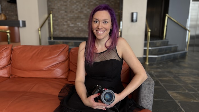 Jessy Poulin pose sur un divan dans le hall d'entrée chic d'un bâtiment. Elle tient une caméra professionnelle dans ses mains.