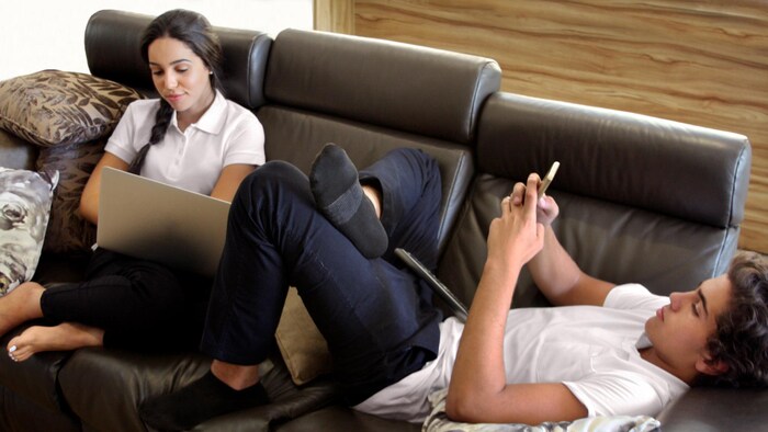 Des adolescents sur un sofa regardent des appareils électroniques.