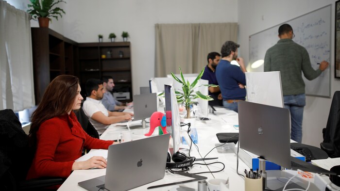 Trois personnes travaillent devant un tableau, alors que trois autres sont assises à l'ordinateur dans une salle avec, en fond, une bibliothèque et des plantes.