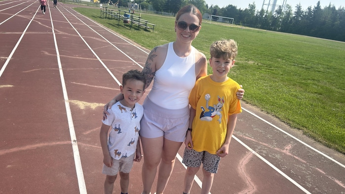 Une jeune femme et deux enfants se tiennent souriant sur une piste d'athlétisme extérieur.