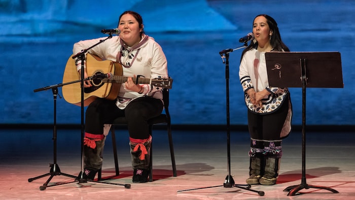 Une jeune fille joue de la guitare et une autre chante à côté d'elle sur une scène.