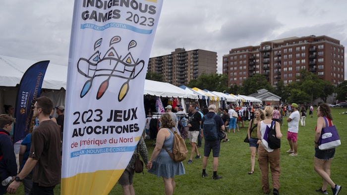 Une bannière avec le logo des Jeux autochtones de 2023 et une foule sur la pelouse près de chapiteaux.