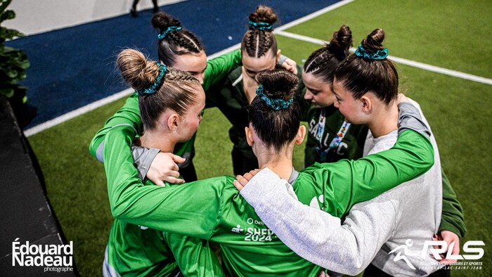 Six jeunes athlètes féminines, portant des uniformes verts et des chignons assortis, se tiennent en cercle dans un vestiaire ou gymnase. Elles sont regroupées en signe de solidarité.