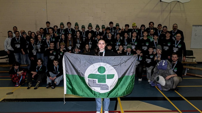 Une grande équipe sportive, vêtue de chandails noirs identifiés « Saguenay-Lac-Saint-Jean », pose dans un gymnase. Au premier plan, une jeune femme tient un drapeau vert et gris orné d'un logo blanc. Plusieurs membres portent des médailles et une mascotte grise est présente.