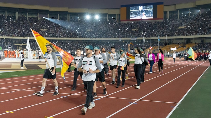 Une douzaine de personnes défilent sur la piste de course du stade, derrière un homme qui porte et agite le drapeau du Nouveau-Brunswick.