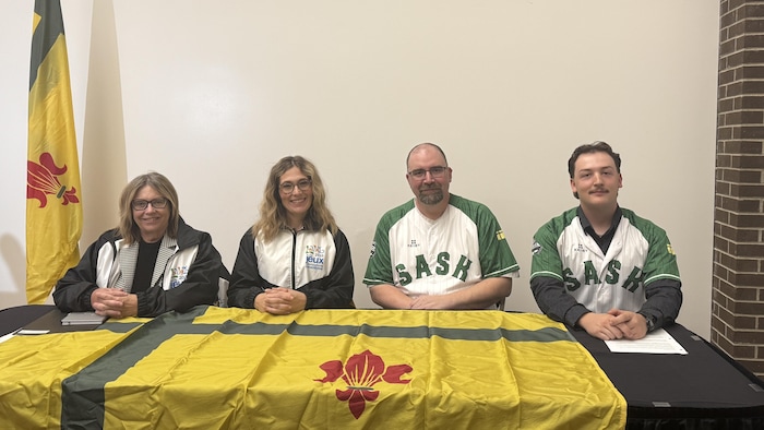 Une photo d'Alana Ross, de Sarah Turnbull, de Denis Simard et de Nathanael Olivier derrière une table où un drapeau de la Fransaskoisie est placé