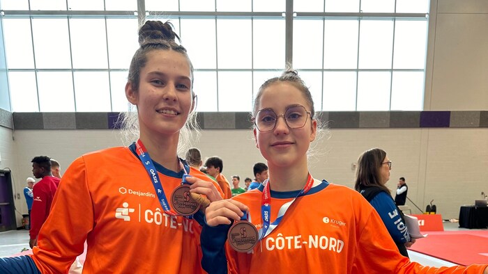 Deux jeunes filles tiennent un drapeau de la délégation de la Côte-Nord et montrent leur médaille de bronze à la caméra. 