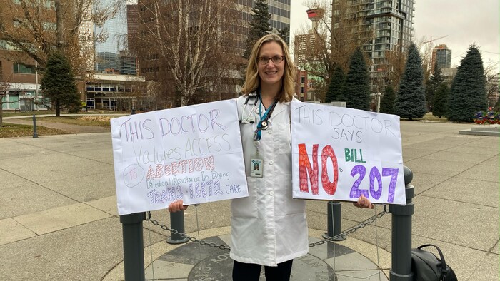 Une dame à l'extérieur avec un sarrau tenant deux pancartes sur lesquelles il est écrit : cette médecin accorde de l'importance à l'accès à l'avortement et cette médecin dit non au projet de loi 207.