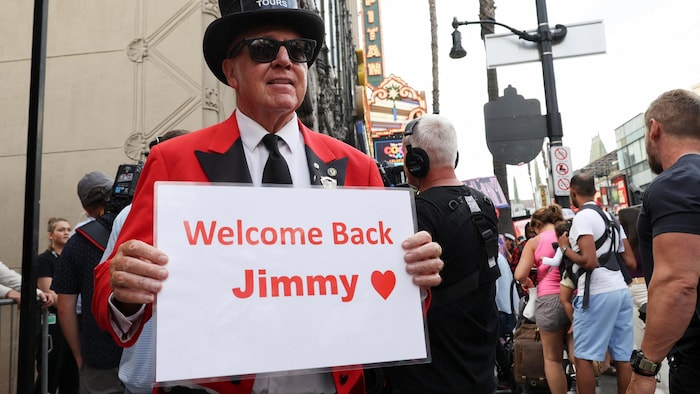 Un homme portant un chapeau noir et une veste rouge tient une pancarte avec le message « Welcome back Jimmy ».