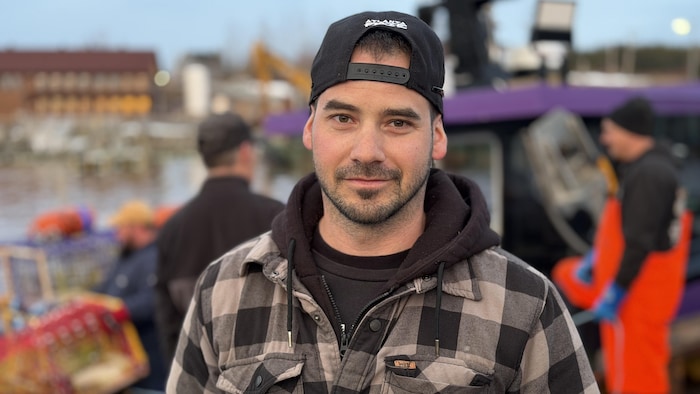 Un portrait du pêcheur Jimmy-Lee Bourget devant le quai de L'Anse-à-Beaufils animé en arrière plan.