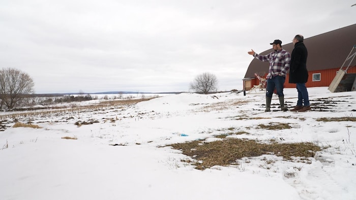 L'agriculteur Joël Laurin et le journaliste Christian Noël discutent dans un champ enneigé près d'une grange rouge, l'un pointant l'horizon du doigt.