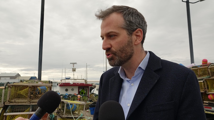 Le ministre est sur un quai avec des casiers à homard et parle derrière plusieurs micros.