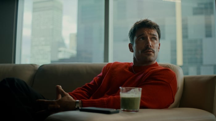 Le personnage est assis dans un condo du centre-ville et regarde une autre personne, un café devant lui.