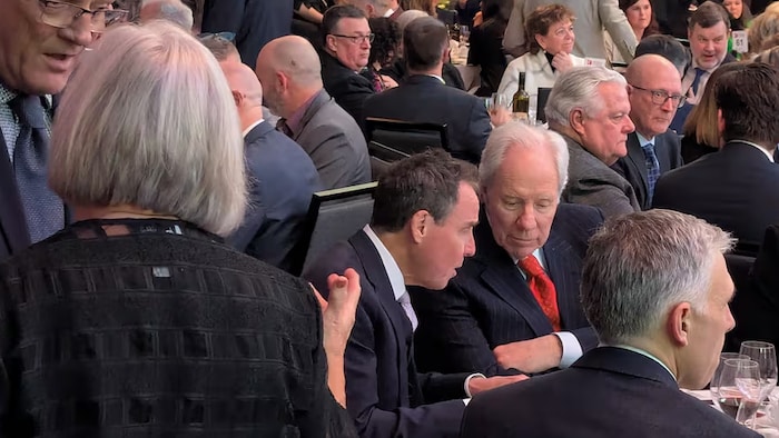 John Herron et Jim Irving sont assis autour d'une table avec d'autres personnes au milieu d'une foule dans une salle de conférence.