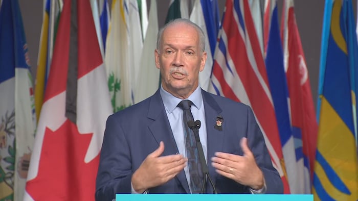 جون هورغان متحدثاً في مؤتمر صحفي، يقف خلف منبر وخلفه علم كندا وأعلام المقاطعات الكندية.