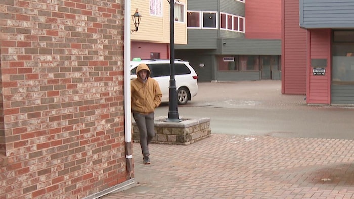 Un homme de loin, les mains dans les poches de son blouson brun clair et un capuchon rabattu sur la tête, marche et arrive près d'un mur de briques.