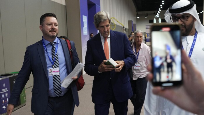 John Kerry marche dans un couloir pendant que quelqu'un le prend en photo sur un téléphone cellulaire.