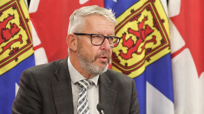 John Lohr, en complet sombre et cravate et chemise claires, est en train de parler en conférence de presse, assis devant des drapeaux de la Nouvelle-Écosse et du Canada.