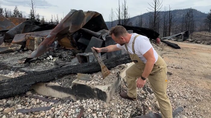 John Rousseau est penché en avant et saisit un morceau de métal carbonisé au milieu des ruines calcinées de son atelier, situé sur sa propriété.