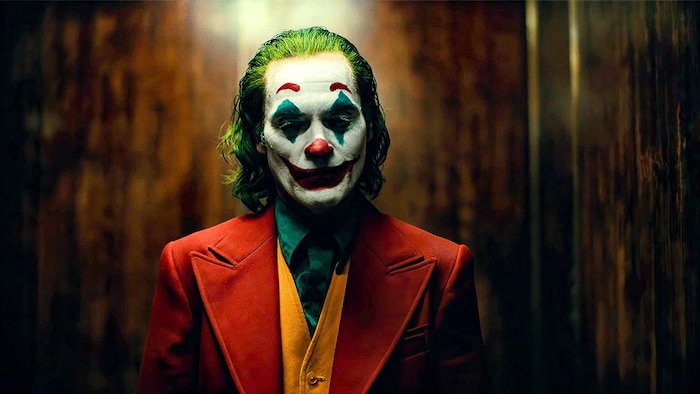 Un homme vêtu d'un veston rouge a le visage maquillé à la manière d'un clown. Ses cheveux sont teints en vert.