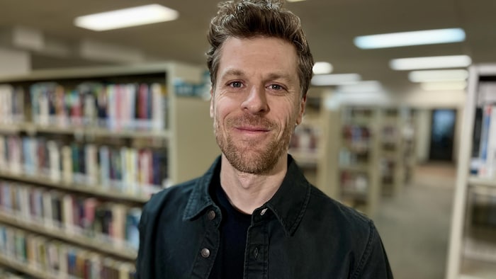 Un homme debout dans une bibliothèque sourit à la caméra.