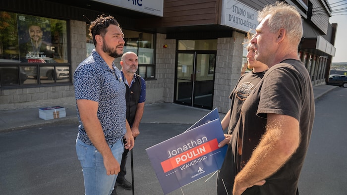 Deux hommes tiennent des pancartes dans leurs mains et discutent avec deux autres hommes devant eux dans un stationnement.