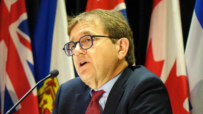 وزير الموارد الطبيعية الكندي جوناثان ويلكينسون متحدثاً في مؤتمر صحفي، أمامه ميكروفون وخلفه علم كندا وأعلام المقاطعات الكندية.