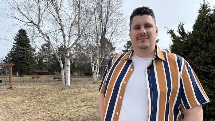 Un homme pose pour la caméra, dehors tôt au printemps. Des arbres dénudés sont visibles derrière.
