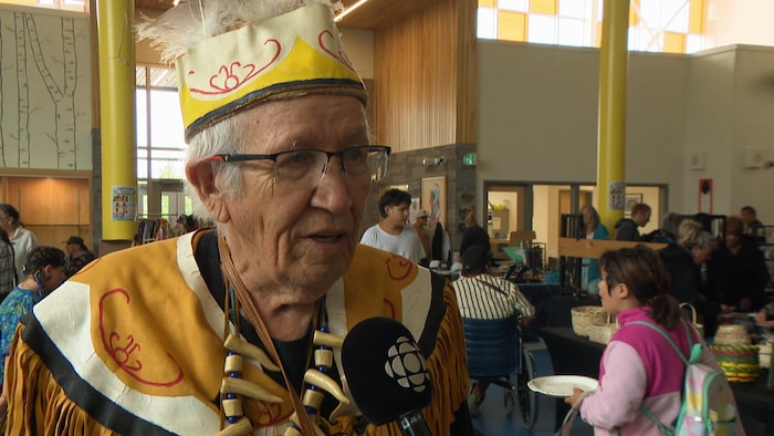 Un homme âgé interviewé au milieu d'une petite foule dans un centre communautaire.