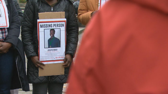 Affiche où il est écrit « personne disparue » avec une photo de Joseph Maku au centre. L'affiche est portée par une femme habillée d'un manteau et entourée d'autres personnes portant eux aussi une copie de l'affiche.