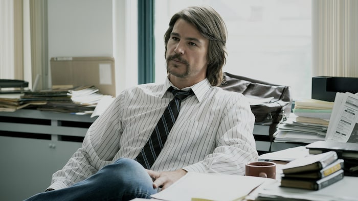 Un homme avec les cheveux mi-longs, vêtu d'une chemise lignée et d'une cravate bleue, est assis à son bureau.