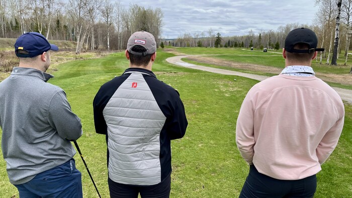 Trois joueurs de golf regardent le terrain à Baie-Comeau, bâtons à la main.