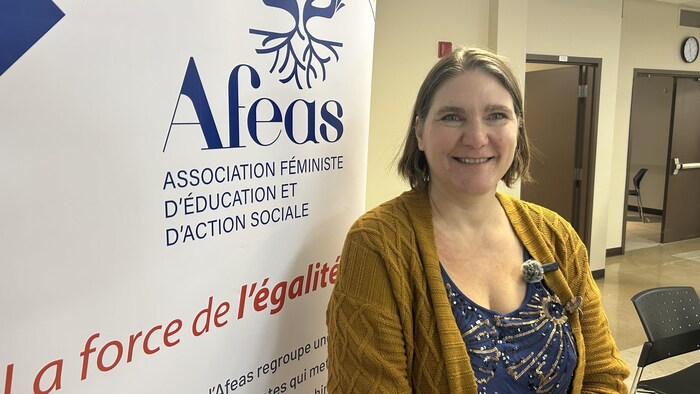 Une femme souriante aux cheveux bruns porte un cardigan moutarde sur un haut bleu pailleté. Elle se tient devant une bannière de l'Afeas. À l'arrière-plan se trouve un couloir d'édifice avec des portes ouvertes et des chaises.