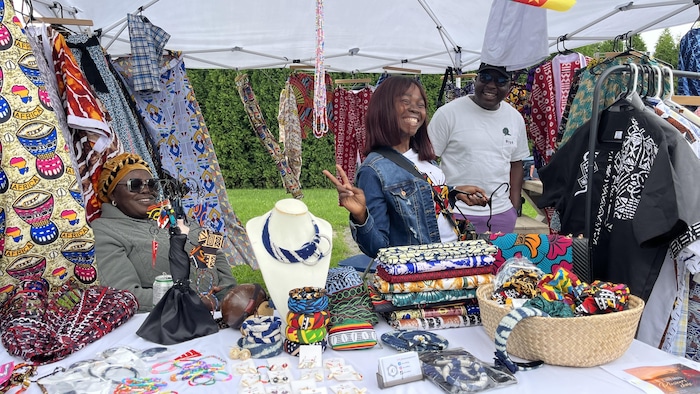 Trois personnes se tiennent sous une tente d'exposition en plein air entourées d'articles artisanaux. La table présente des bijoux, des tissus pliés et des accessoires colorés, tandis que des vêtements imprimés sont suspendus autour d'elles.