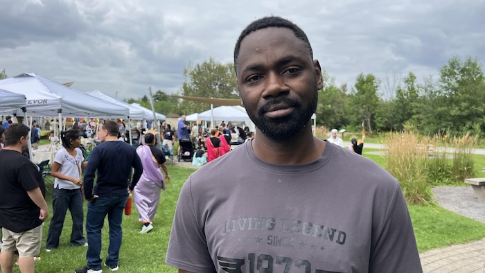 Un homme noir barbu regarde la caméra en gros plan. En arrière-plan, sous un ciel nuageux, des personnes se rassemblent autour de tentes dans un parc herbeux.