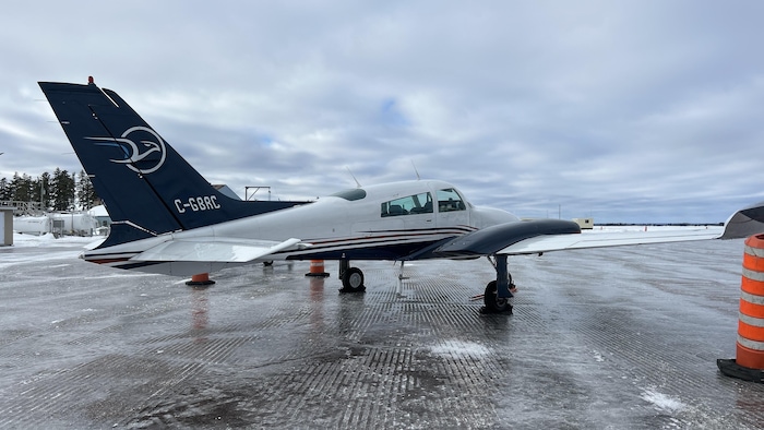 Cette photo présente un avion bimoteur blanc et bleu, immatriculé C-GBRC, stationné sur une piste glacée. L'appareil est entouré de cônes de signalisation orange sous un ciel couvert, avec des installations aéroportuaires et une forêt en arrière-plan.