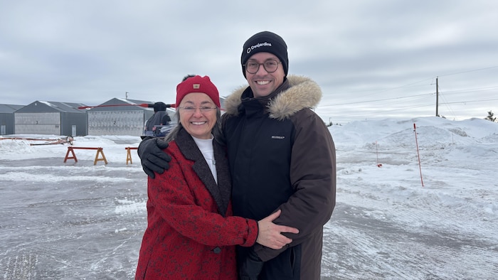 Un homme et une femme souriants posent ensemble à l'extérieur en hiver. Ils portent des vêtements chauds, des tuques et des gants. Derrière eux se trouvent des hangars d'aviation et d'importants bancs de neige sous un ciel gris.