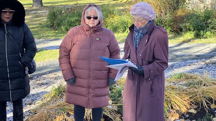 Une dame, habillée en hiver, lit un texte lors d'une cérémonie près de deux autres femmes.