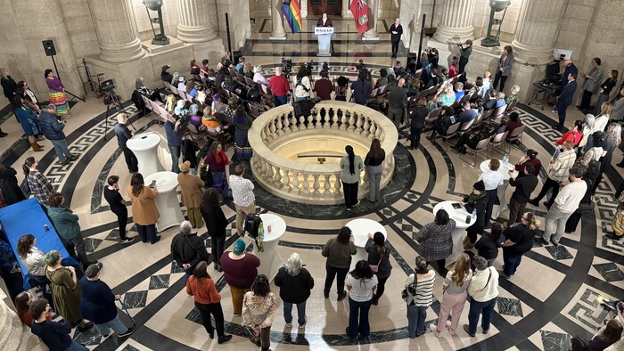 Une foule dans le Palais législatif du Manitoba debout ett assises devant une personne  qui fait un discours à un podium, flanquée du drapeau de la province et du drapeau 2ELGBTQQIA+.