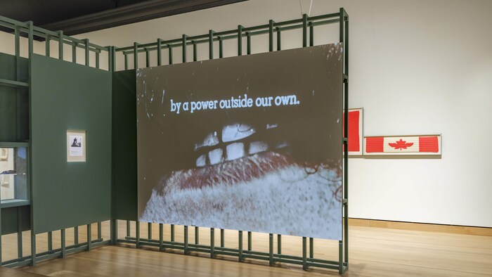 Un film qui montre la bouche d'un homme est projeté dans un musée devant une œuvre qui représente le drapeau du Canada.