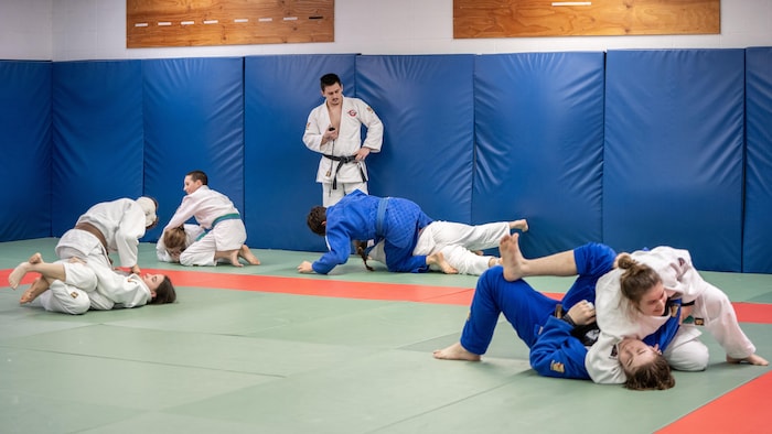 Un entraîneur, debout, tient un chronomètre pendant que plusieurs duos de judokas s’entraînent au sol sur les tatamis d’un dojo.