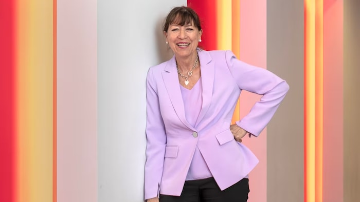 Jodi Cameron wears a pink shirt and smiles as she leans against a wall.