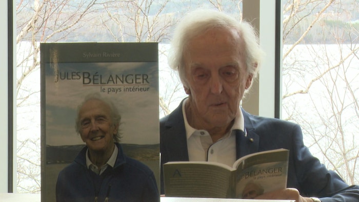 Jules Bélanger lit la biographie écrite par Sylvain Rivière devant une fenêtre.