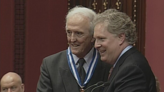 Jules Bélanger et Jean Charest se serrent la main et sourient à des photographes.