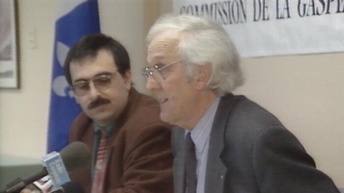 Assis à une table, Jules Bélanger lit un texte dans un micro posé sur une table.