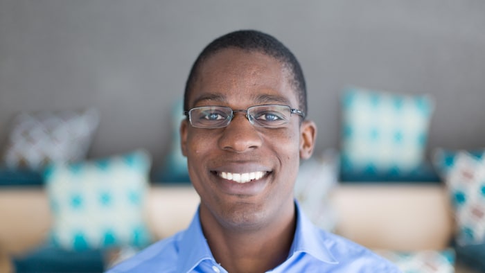 A smiling man in glasses poses in front of wooden benches and bright pillows. 