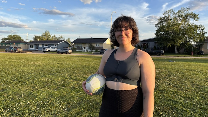 Une jeune femme avec des lunettes et des cheveux courts se tient debout sur un terrain d'herbe en fin de journée, tenant un ballon de rugby.