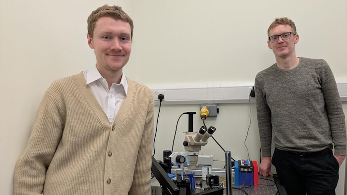 Deux hommes grands et minces sont debout autour d'une table où est posé un microscope. Ils se tournent vers la caméra pour la photo.